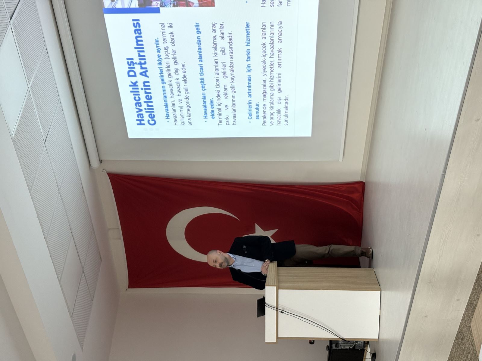 Ahmet Orçun Songur is seen delivering a presentation at the lectern. In the background, there is a Turkish flag and projected presentation slides. The slides feature titles such as "Increasing Non-Aeronautical Revenues" and "Development of Airport Commercial Strategies
